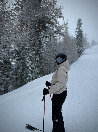 A skier dressed in winter gear stands on a snowy slope, holding ski poles and looking ahead. The background features snow-covered trees and a misty, overcast sky, evoking a serene winter atmosphere. The skier's helmet prominently displays the "SNÖVIA" logo, emphasising stylish and functional equipment for snowy adventures.